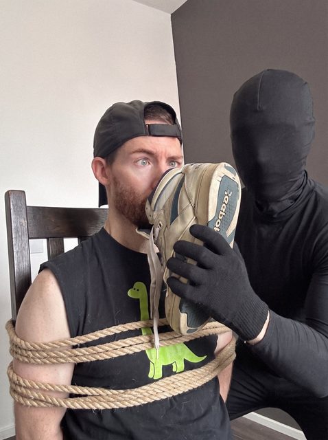 A man tied to a chair has a sneaker held to his face by another man in a full black suit and gloves.