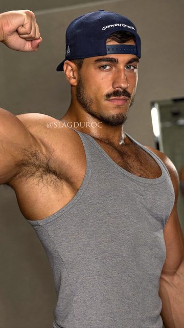 A muscular man in a tank top and cap flexes his arm.