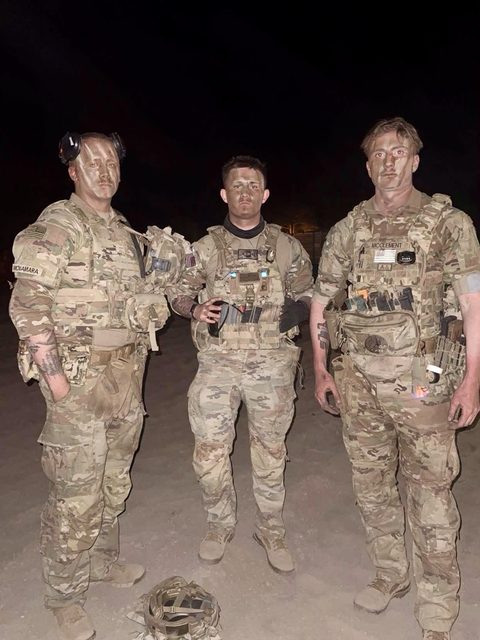 Three men in full military camouflage uniforms and tactical gear stand outdoors at night.
