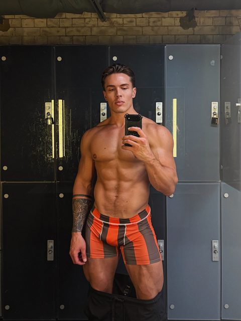 A muscular man takes a mirror selfie in a locker room wearing striped boxer briefs.
