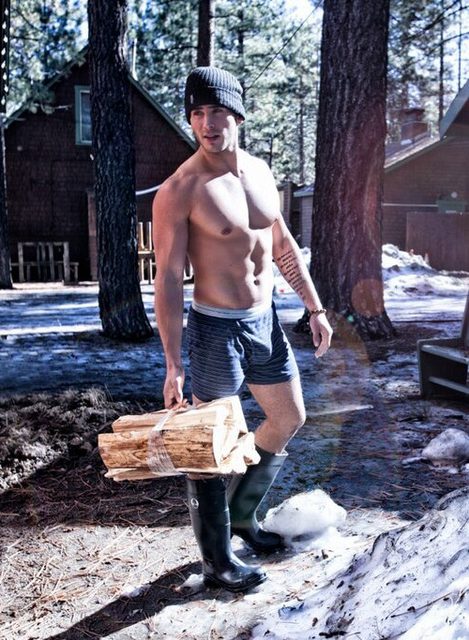 A muscular man in a beanie and boxer shorts carries firewood through a snowy outdoor setting.