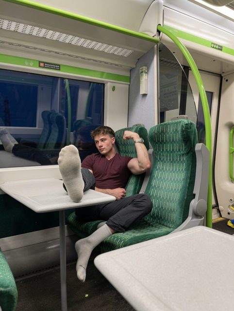 A muscular man flexes his arm while relaxing on a train with his feet up on a table.