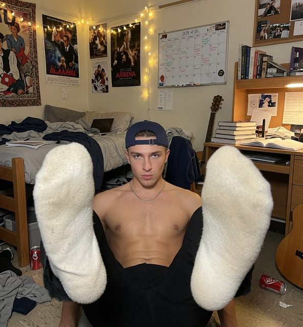 A shirtless man in a cap sits on the floor of his bedroom, displaying his sock-covered feet.
