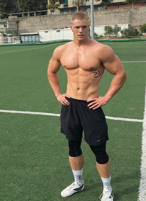 A muscular man in sportswear stands on an outdoor sports field.