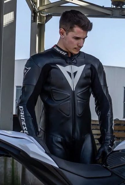 A young man wearing a full black and white Dainese leather motorcycle suit and matching gloves stands outdoors next to a bike.