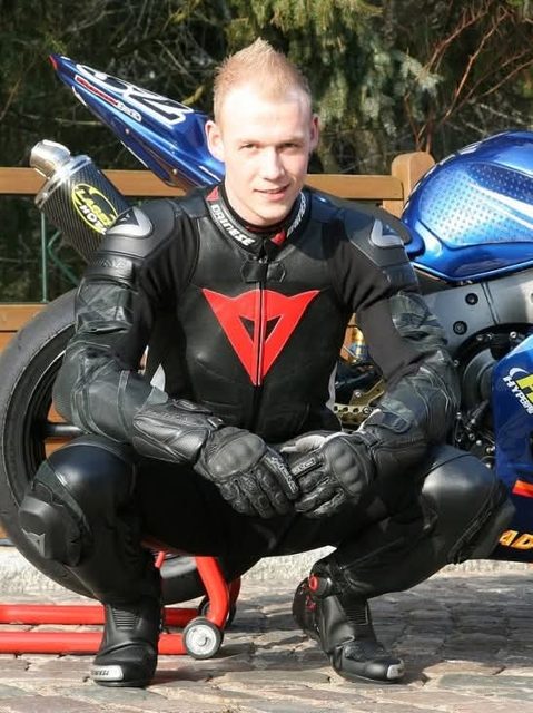 A man in a full black and red Dainese leather motorcycle suit squats in front of a blue sportbike outdoors.