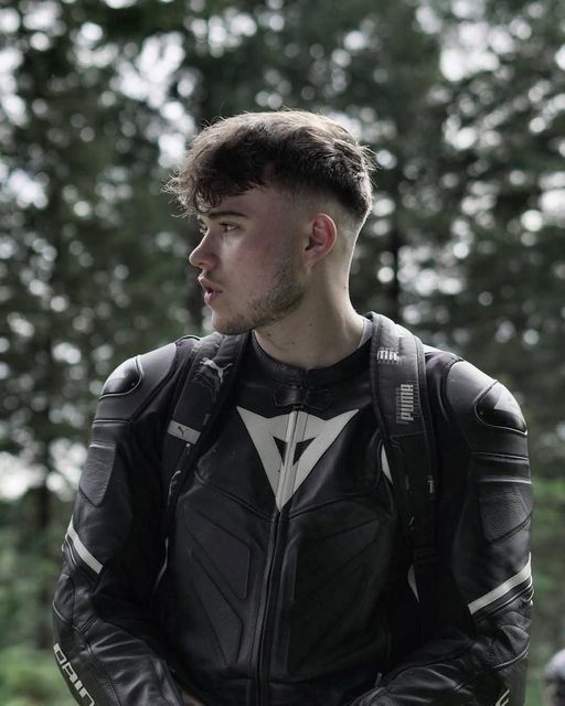 A young man wearing a black leather motorcycle jacket and backpack looks off to the side outdoors.