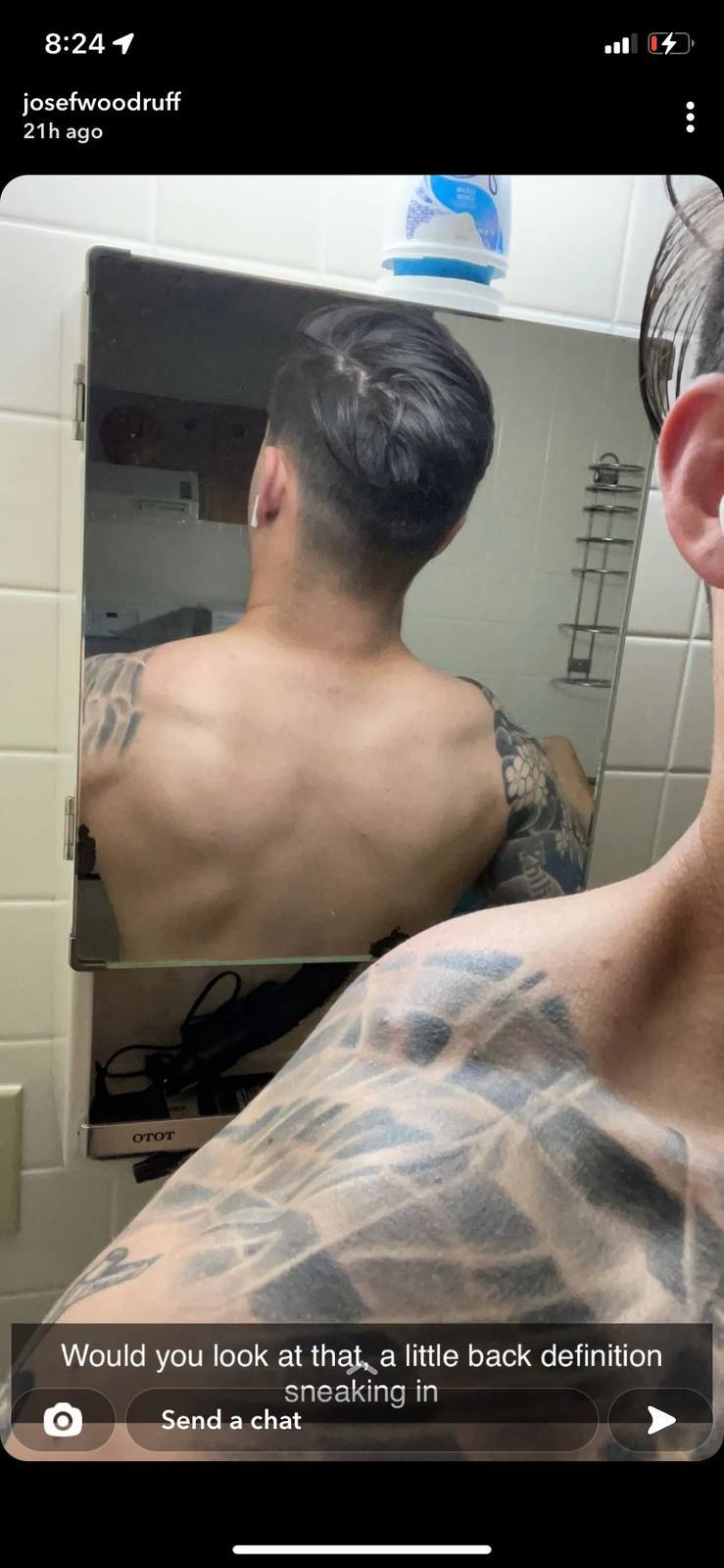 A man with tattoos is seen from behind, reflected in a bathroom mirror.