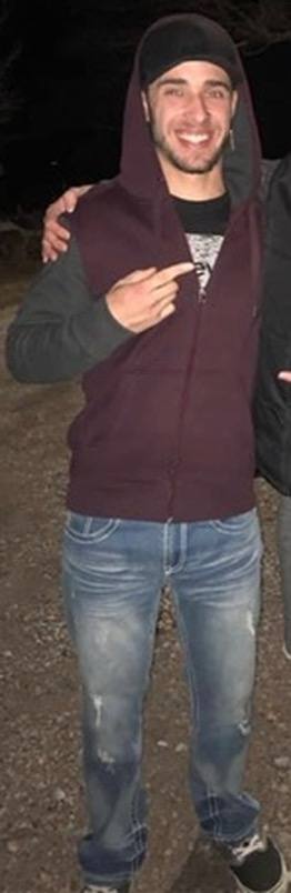 A man with a beard, cap, and denim jeans smiles outdoors at night.
