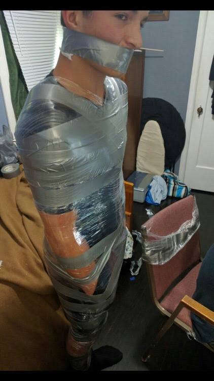 A young man is fully restrained with tape, including a mouth gag, in an indoor setting.