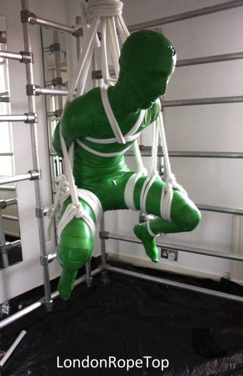 A person in a green full body suit and mask is suspended by white ropes in an indoor setting.