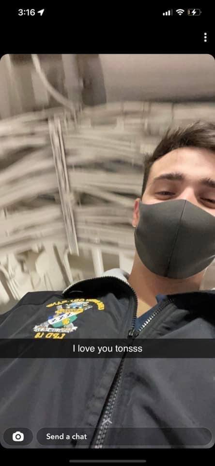 A young man in a naval utility jacket and face mask stands in a ship's interior with visible overhead piping.