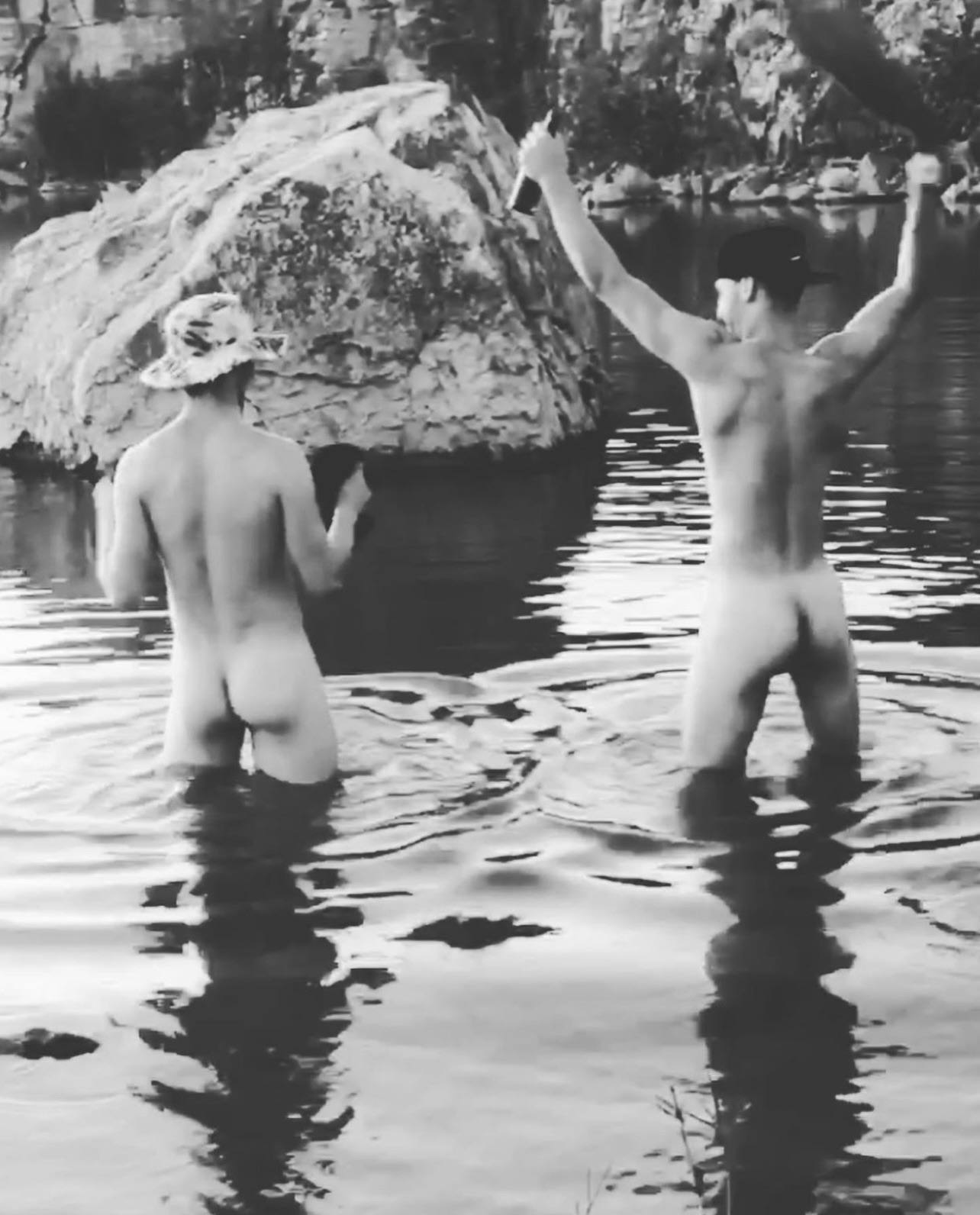 Two naked young men stand in the water of a lake with their backs to the camera, wearing only hats while skinny dipping.