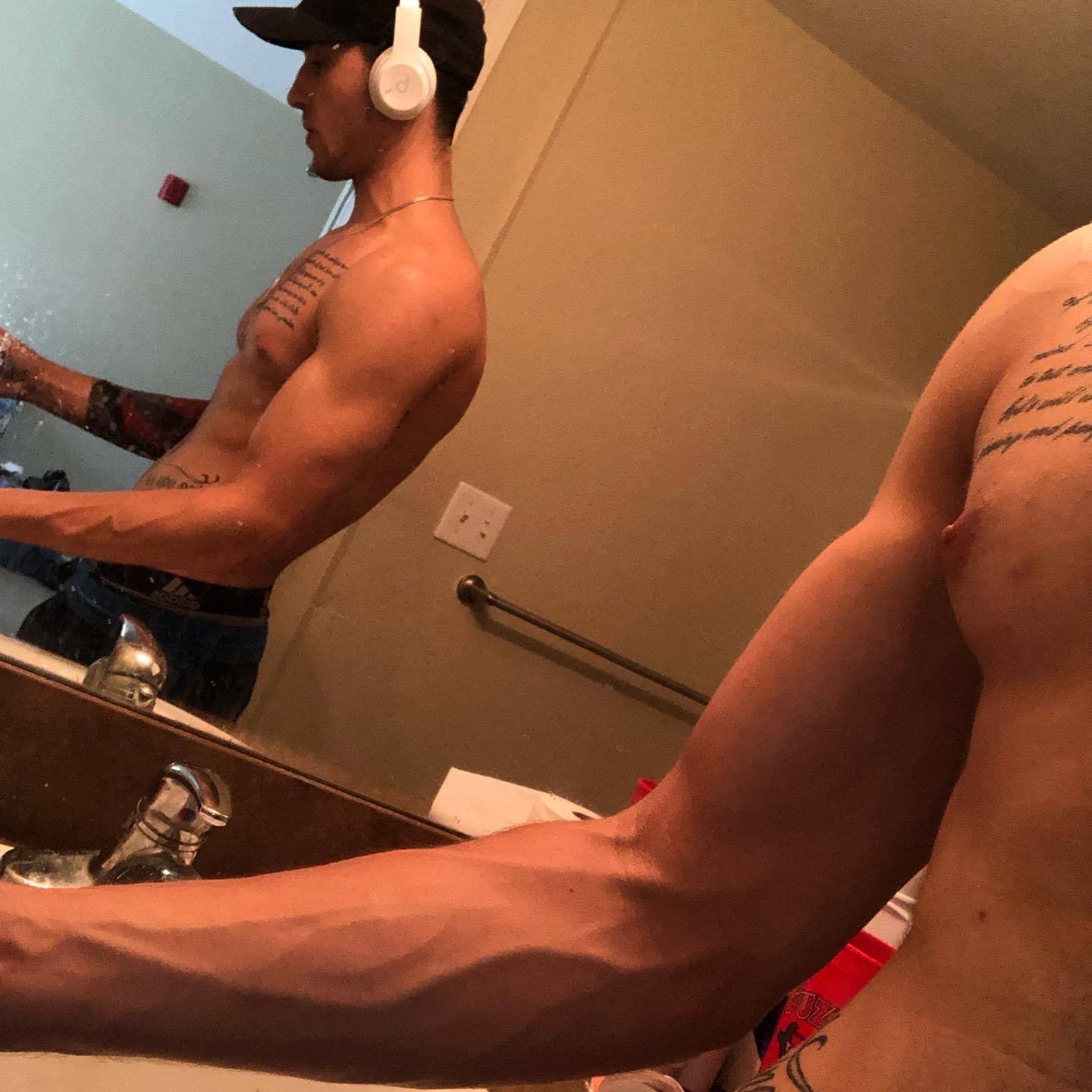 A shirtless, tattooed man takes a mirror selfie in a bathroom, showcasing his vascular arms and muscular build while wearing a baseball cap and headphones.