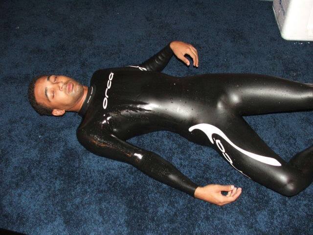 A man in a wet black wetsuit lies on a blue carpet indoors.