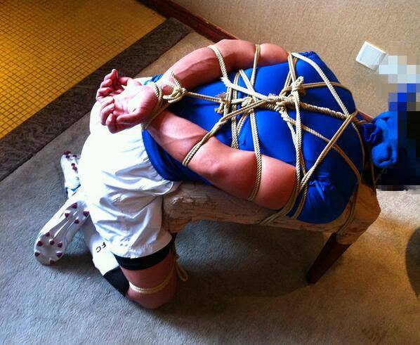 A person in sportswear is secured with rope bondage to a wooden bench.