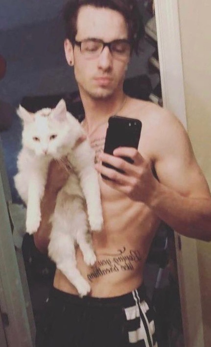 A smooth-bodied man with glasses and tattoos takes a mirror selfie while holding a white cat.