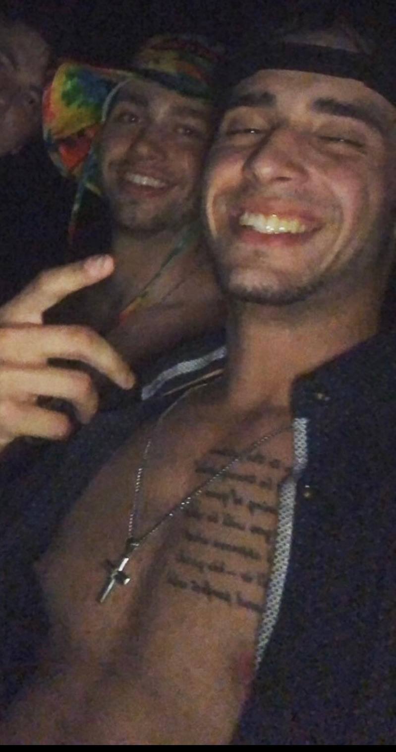 A shirtless twunk with a chest tattoo and a cross necklace smiles for a selfie with friends at night.