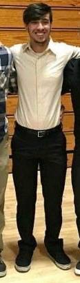 A smiling young man with a beard stands in a gymnasium wearing a beige button-down shirt tucked into black trousers.