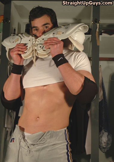 A muscular man poses in a locker room, wearing a cropped shirt and football gear.