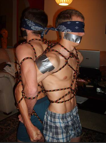Two blindfolded men are tied together back-to-back with rope and gagged with duct tape indoors.