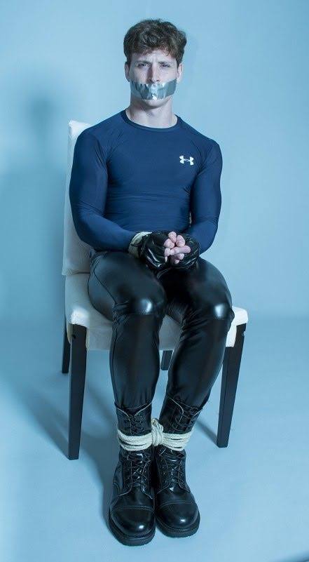 A man in a blue shirt, leather pants, and boots is tied to a chair with rope and gagged with tape.