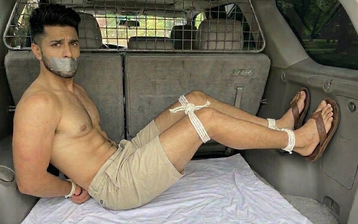 A shirtless man with his mouth taped and limbs bound by rope is restrained in the back of a vehicle.