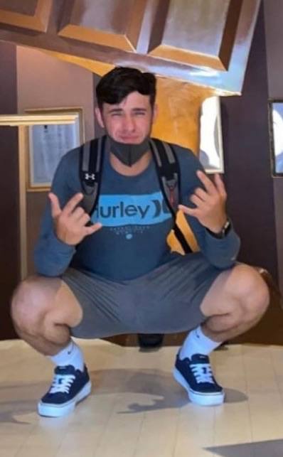 A muscular man in sportswear, wearing a mask and backpack, squats indoors while making hand gestures.
