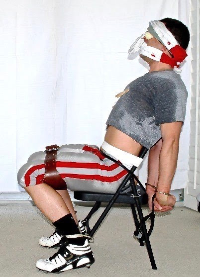 A man is restrained to a chair with a blindfold, gag, handcuffs, and a padded harness.
