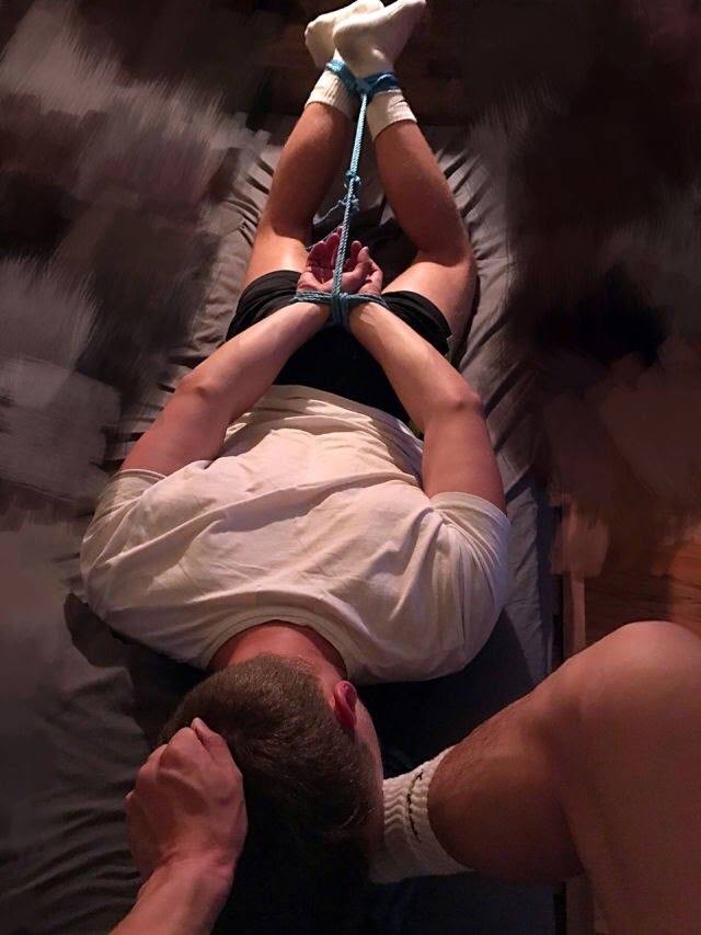 A man is tied with rope on a bed while another man's hand is on his head.