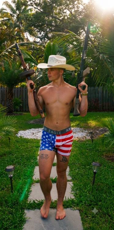 A young man stands shirtless in American flag boxer briefs and a cowboy hat, holding two firearms in a sunny backyard.