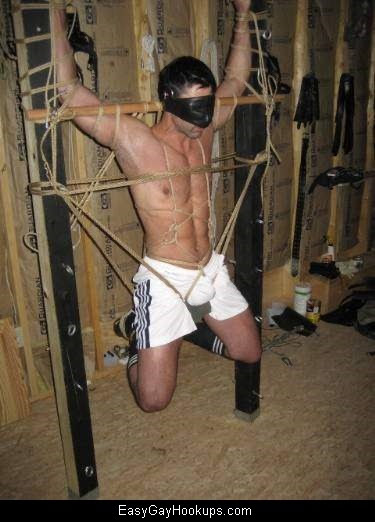 A muscular man is suspended in rope bondage, wearing a leather mask and sportswear.