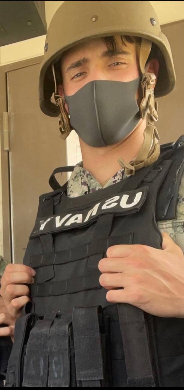 A young man in a military uniform, tactical vest, helmet, and face mask poses indoors.