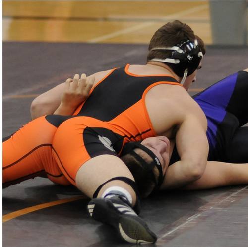 Two muscular men in wrestling singlets grapple on a mat during a match.