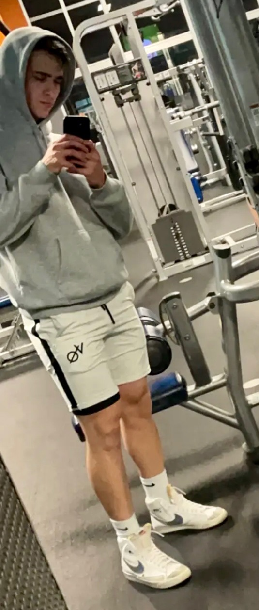 A young man in a hoodie and shorts takes a mirror selfie in a gym.
