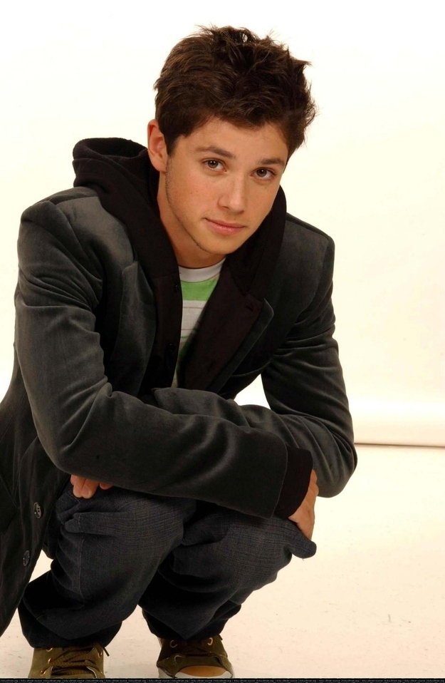 A young man crouches in a studio wearing a jacket, hoodie, and jeans.
