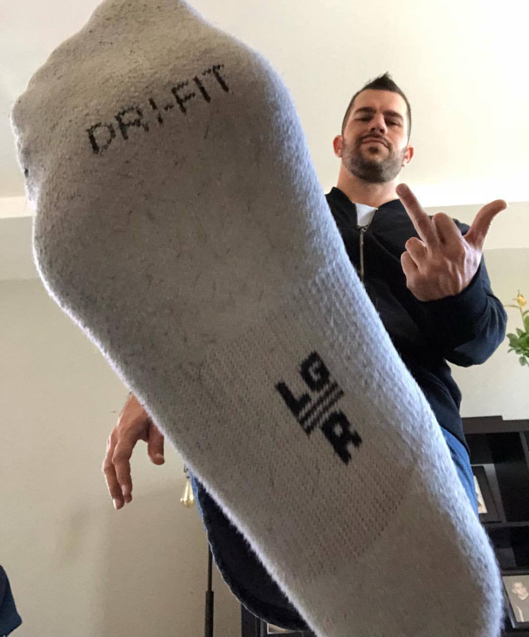 A man in a dark jacket and jeans shows his socked foot and gives the middle finger.