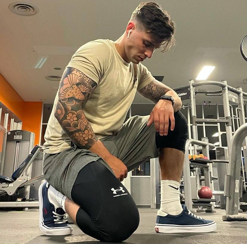 A muscular man with tattoos kneels on the gym floor wearing sportswear and sneakers.