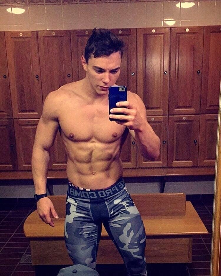 A muscular man takes a mirror selfie in a locker room.