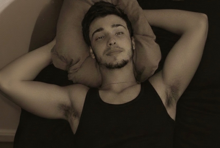 A young man with a light beard and hairy armpits lies on a pillow in a black tank top, looking up at the camera.