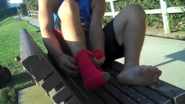 A person sits on an outdoor bench, putting on a pink sock while one foot is bare.
