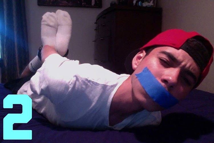 A young man with a red cap is lying prone on a bed, gagged with blue tape and with his feet restrained.