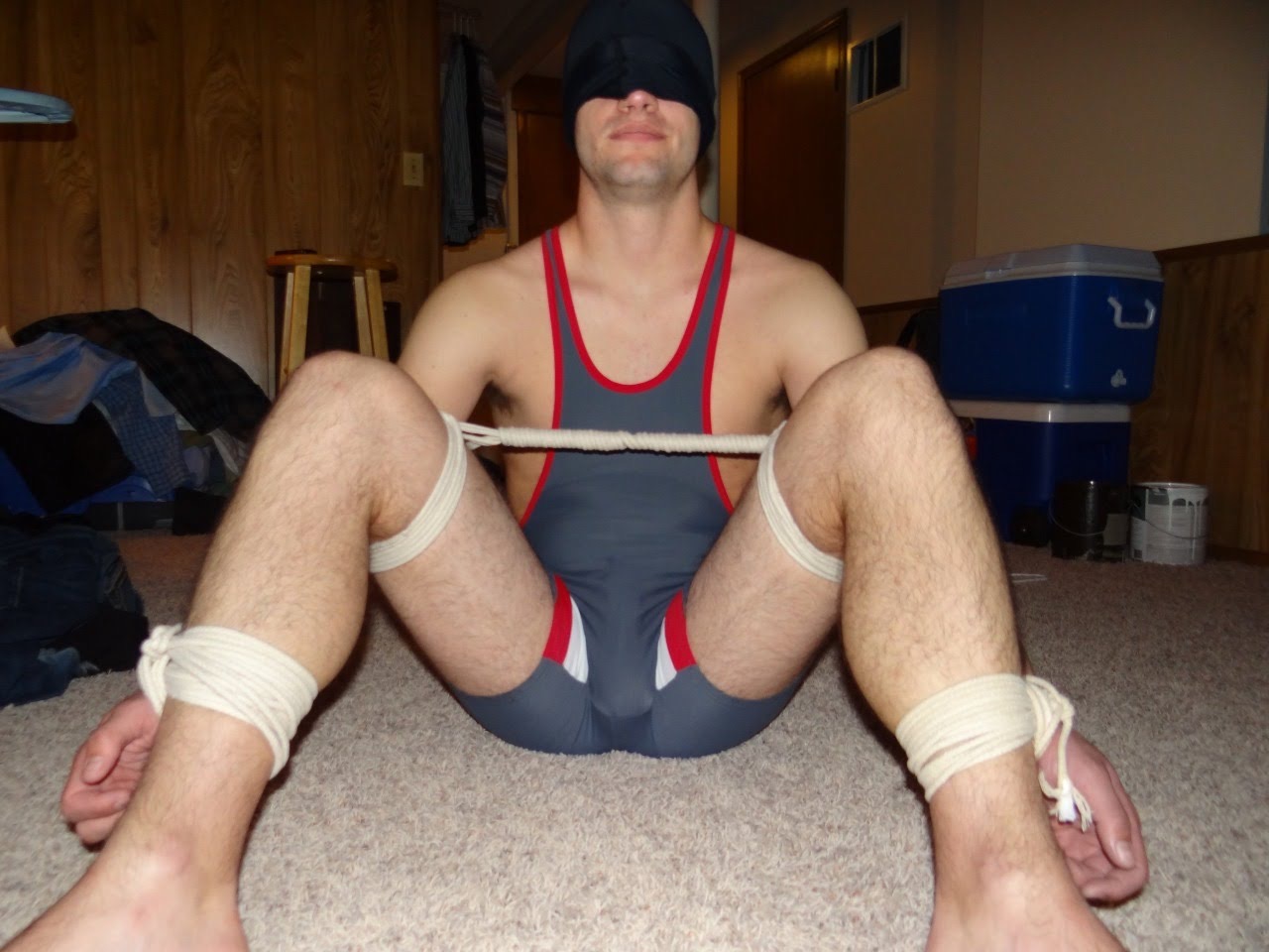 A man wearing a wrestling singlet and blindfold is restrained with rope on the floor.