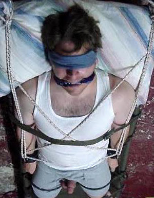 A man in a tank top and shorts is blindfolded and gagged, tied with ropes and straps in a bedroom setting.