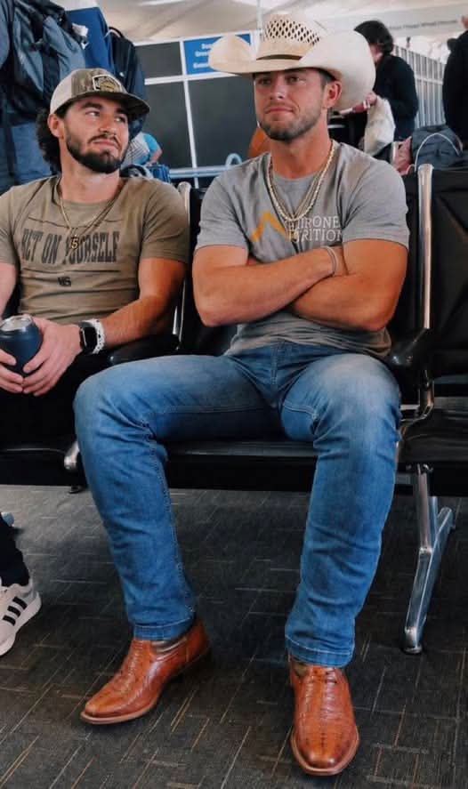 Two masculine men sit in a public waiting area, featuring one man in a cowboy hat and tight denim jeans sitting with his legs spread.