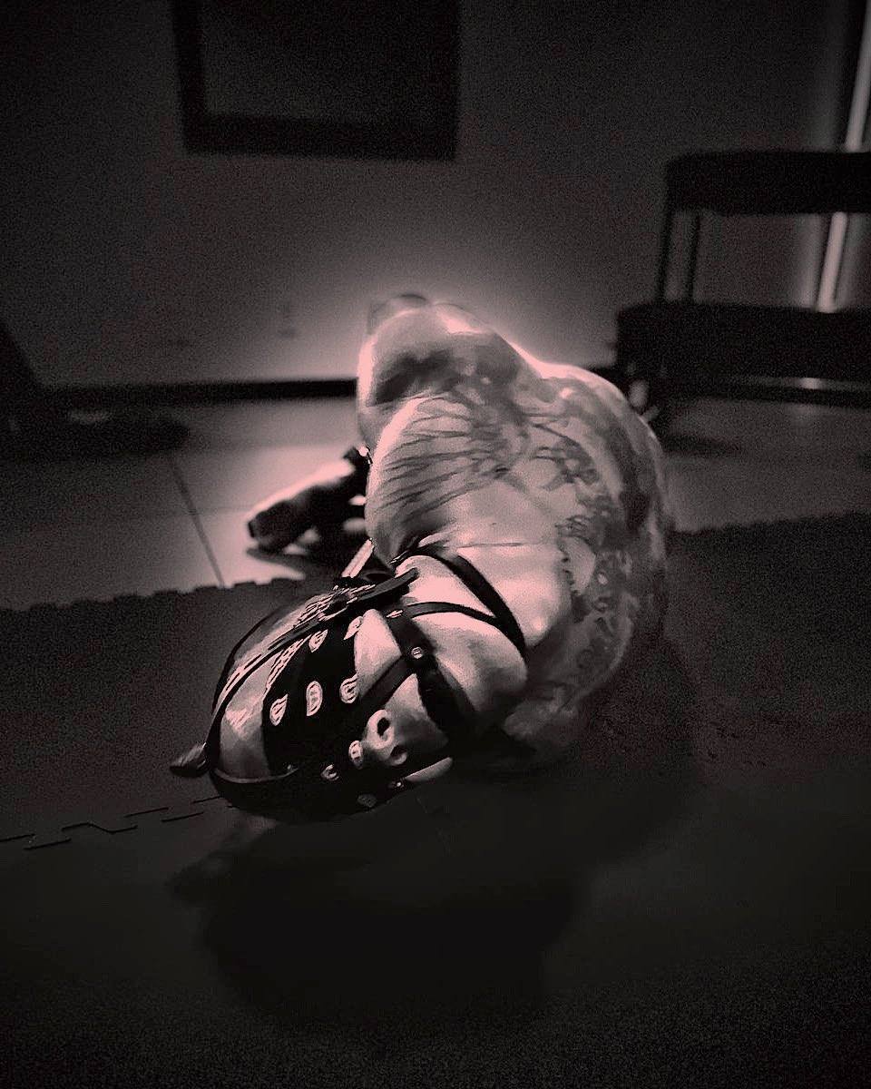 A muscular, tattooed man wearing a leather pup mask and harness leans forward on a padded floor in a dimly lit room.