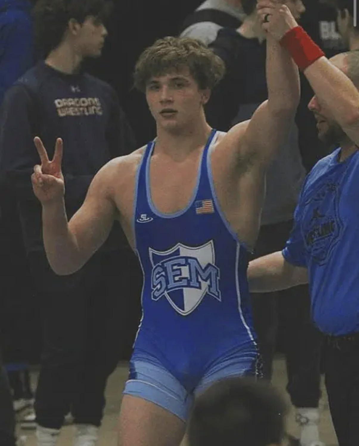 A young muscular man in a blue wrestling singlet makes a victory sign in a gym setting.