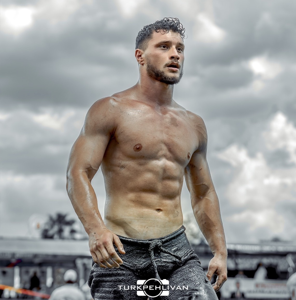 A muscular man wearing traditional wrestling pants stands outdoors under a cloudy sky.