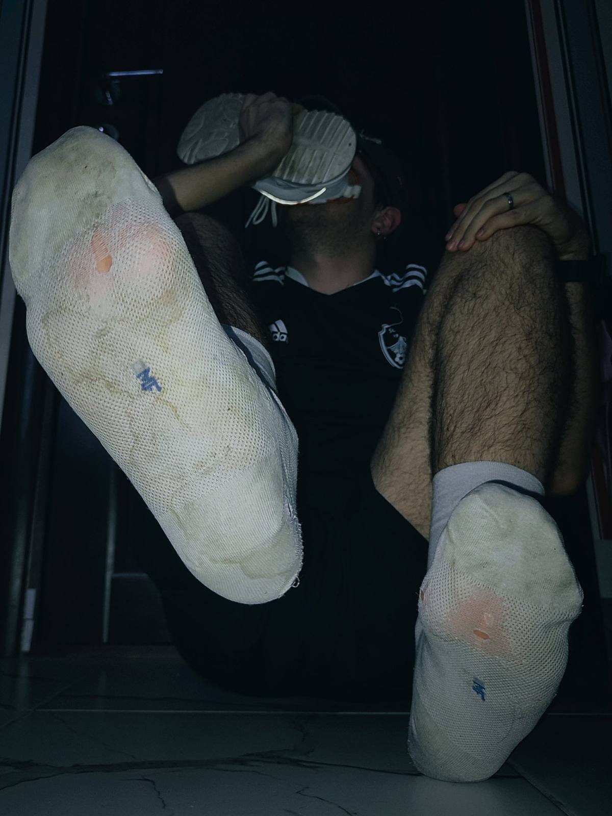 A man holds a sneaker to his face, showing his worn socks and hairy legs.