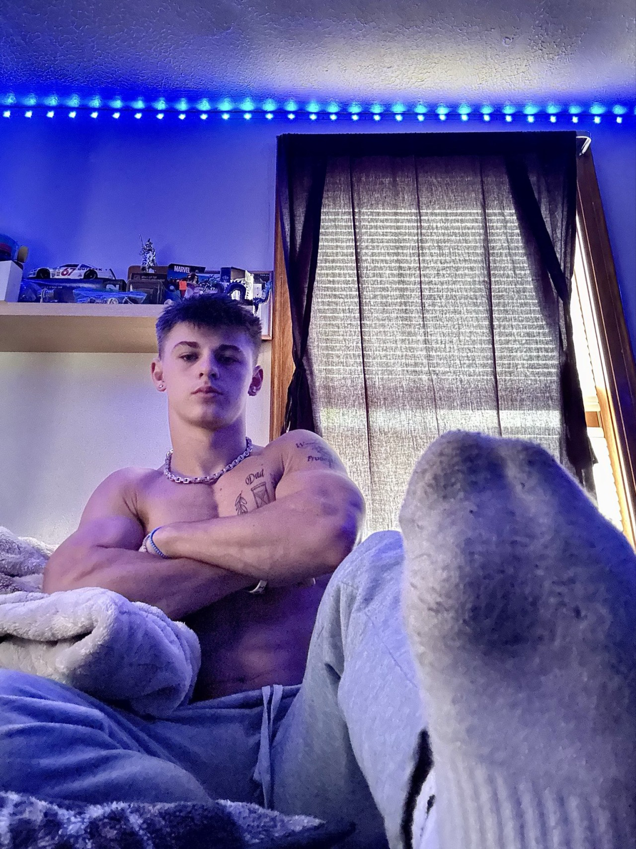 A muscular twink sits in a bedroom with blue LED lighting, wearing sweatpants and socks.